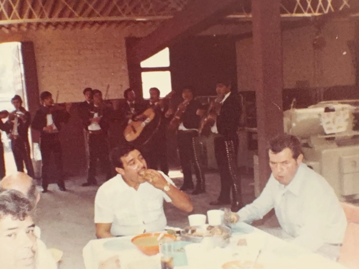 Comida en la nave original de Europlast en los primeros años, con mariachi al fondo; el fundador es el hombre de la derecha de la mesa